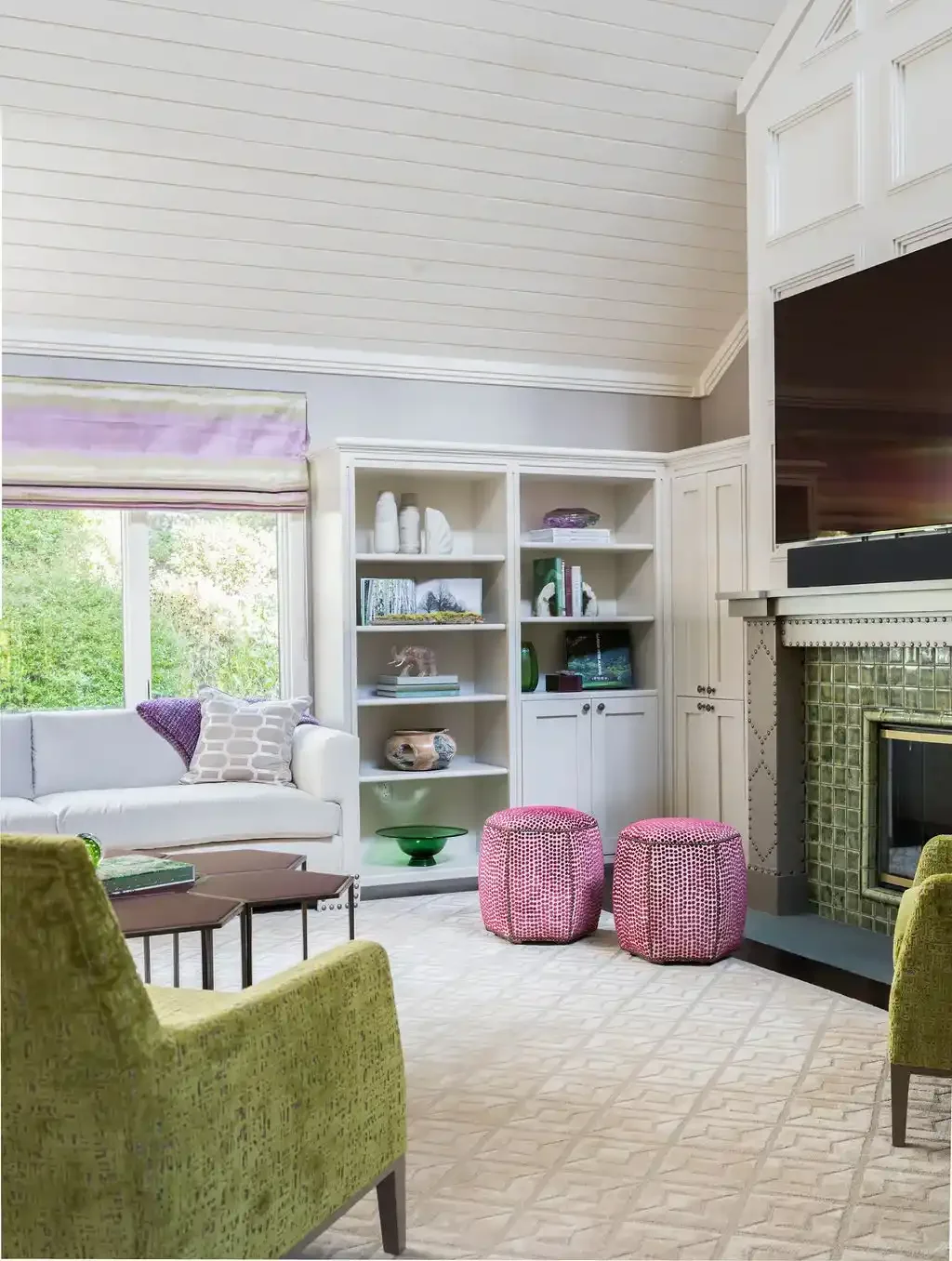 Living room with a white sofa, green armchairs, pink ottomans, built-in white shelves, fireplace with green tiles, and large window with view of trees.
