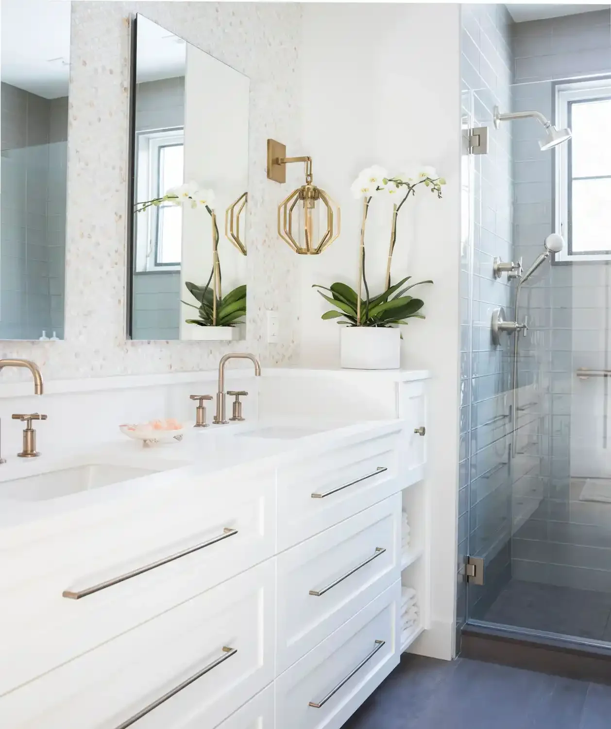 Modern bathroom with white double vanity, gold fixtures, and a walk-in shower with gray tiles and a window.