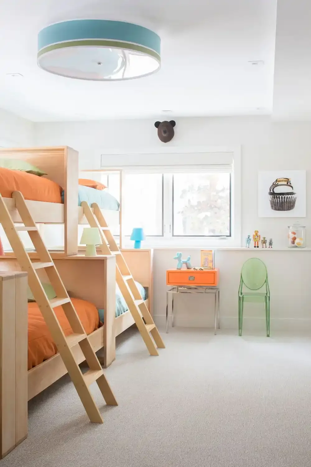 A children's bedroom with a wooden bunk bed that has orange and blue bedding, colorful lamps, stuffed animals, and artwork on the walls. There is a window, a green transparent chair, and a small orange drawer.