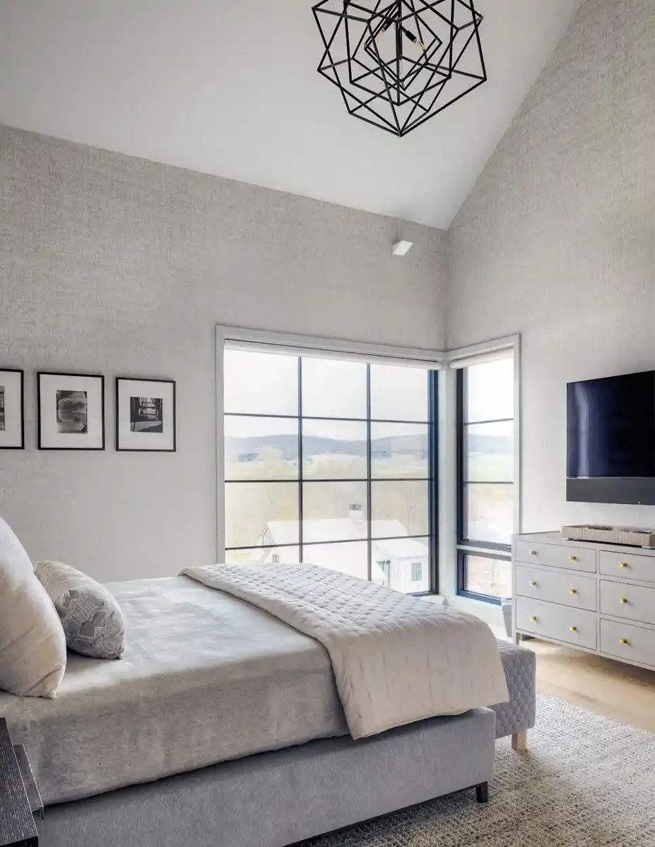 Bright bedroom with large windows, a bed with white bedding and pillows, a white dresser with gold knobs, a wall-mounted TV, black picture frames on the wall, and a modern geometric ceiling light fixture.