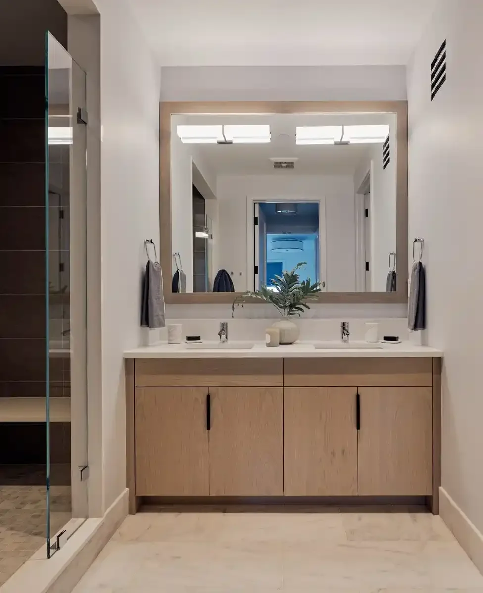 Modern bathroom vanity with a large framed mirror, two sinks, a potted plant, and a white countertop. To the left, a glass shower enclosure and a hallway are visible.