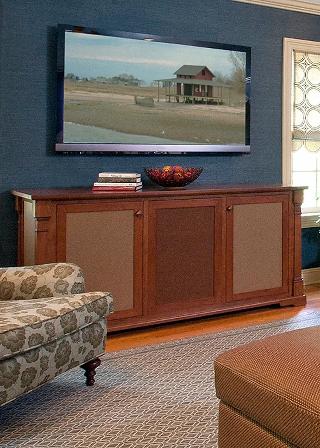 Living room with a flat-screen television mounted on a teal wall, a wooden cabinet below holding a decorative bowl and a stack of books, a floral-patterned sofa, and a window with decorative curtains.