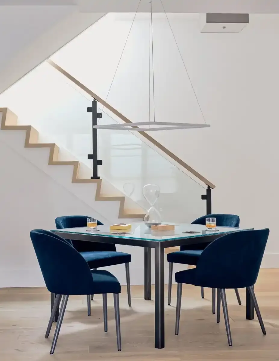 Contemporary dining area with a glass table, four dark blue velvet chairs, a modern pendant light, and a staircase with glass railing in the background.