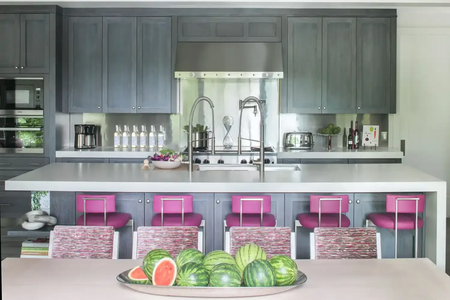 Modern kitchen with dark gray cabinets, white countertop, and a large island with pink chairs. Watermelons are on a tray on the foreground table.