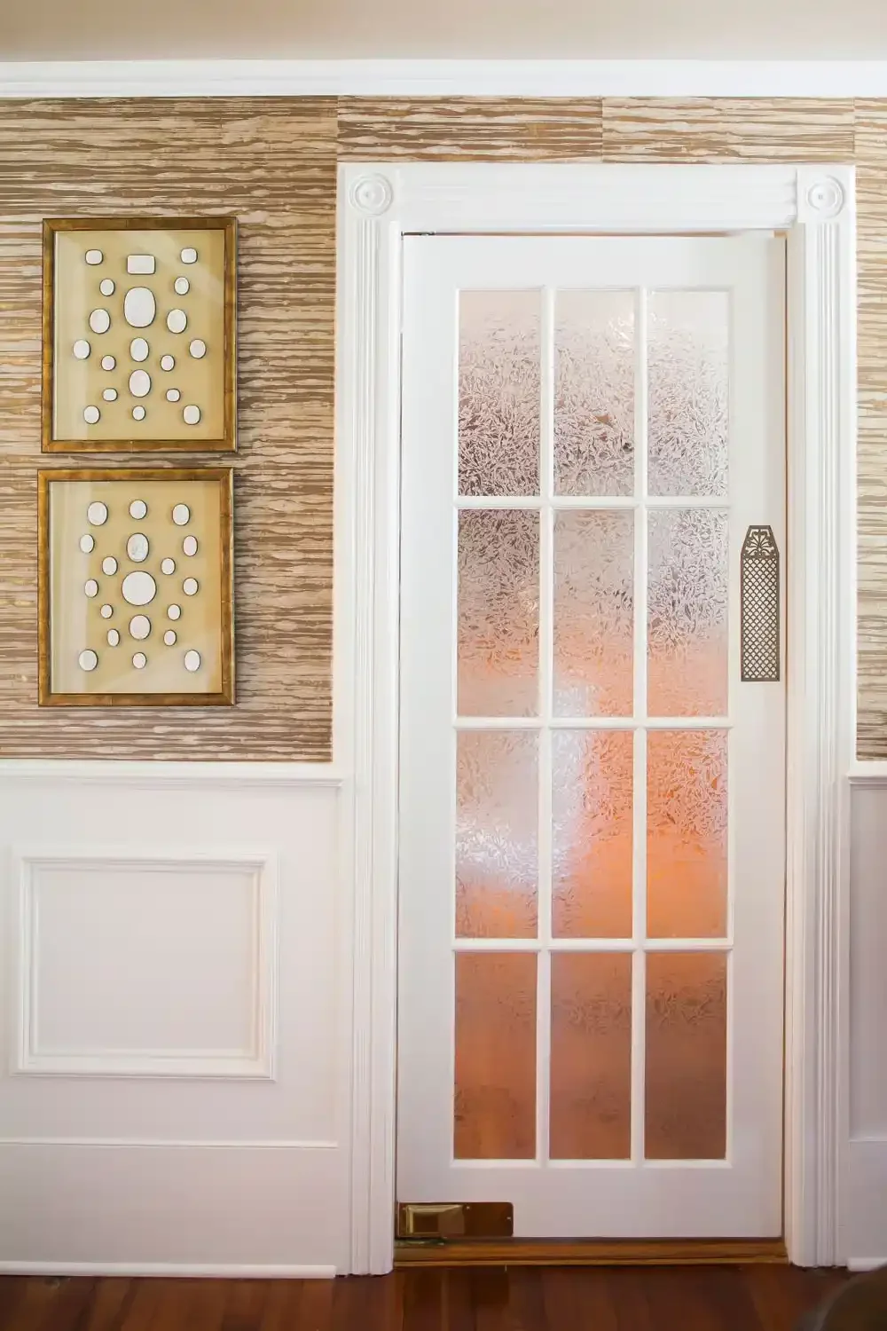 Interior door with frosted glass panels and decorative trim, flanked by wallpapered walls with artwork and wainscoting.