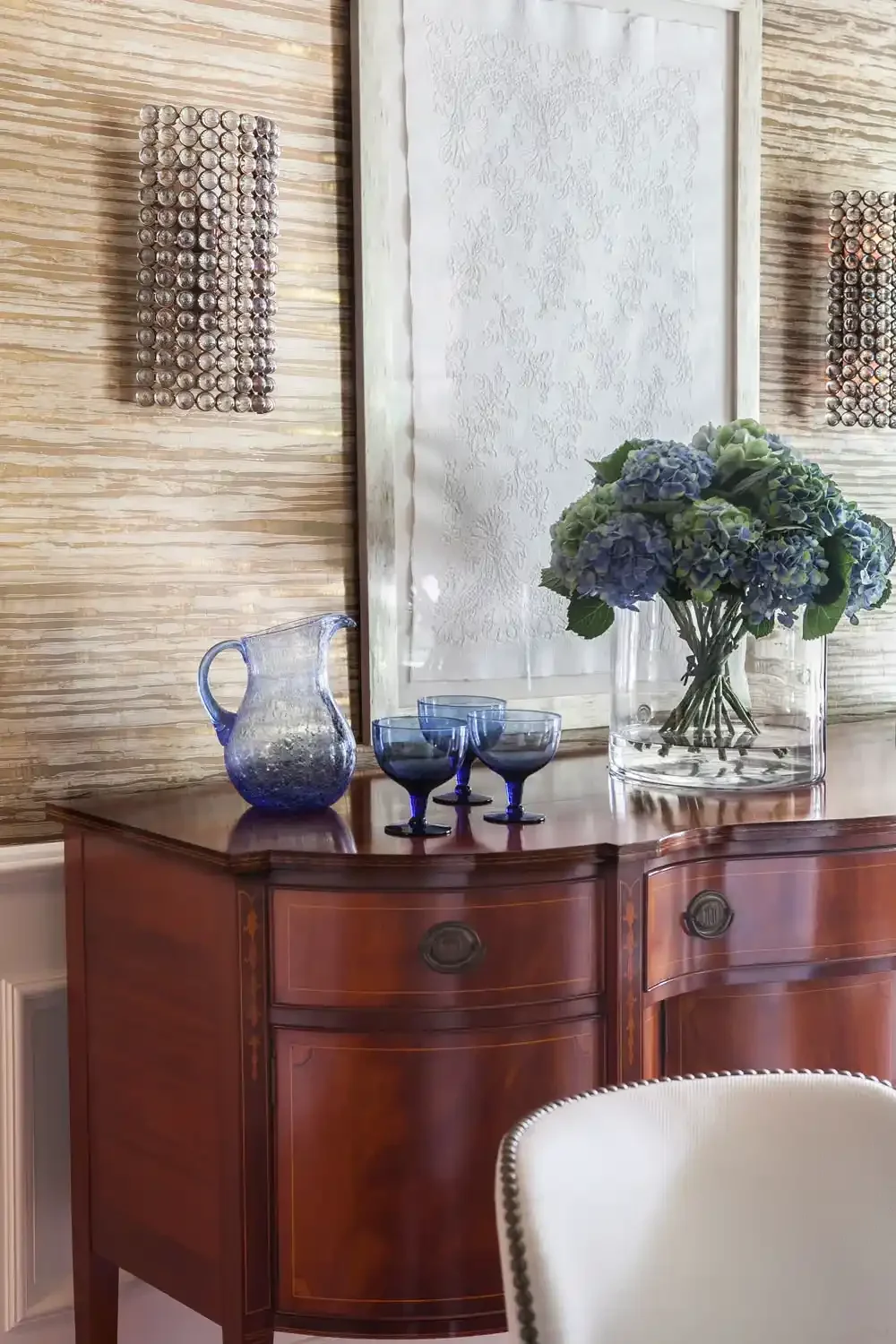 A wooden sideboard with a glass vase of blue and green hydrangeas, a blue glass pitcher and three blue glass goblets on top, against a textured wooden wall with two wall sconces holding decorative metal spheres, and a framed textured artwork in the b
