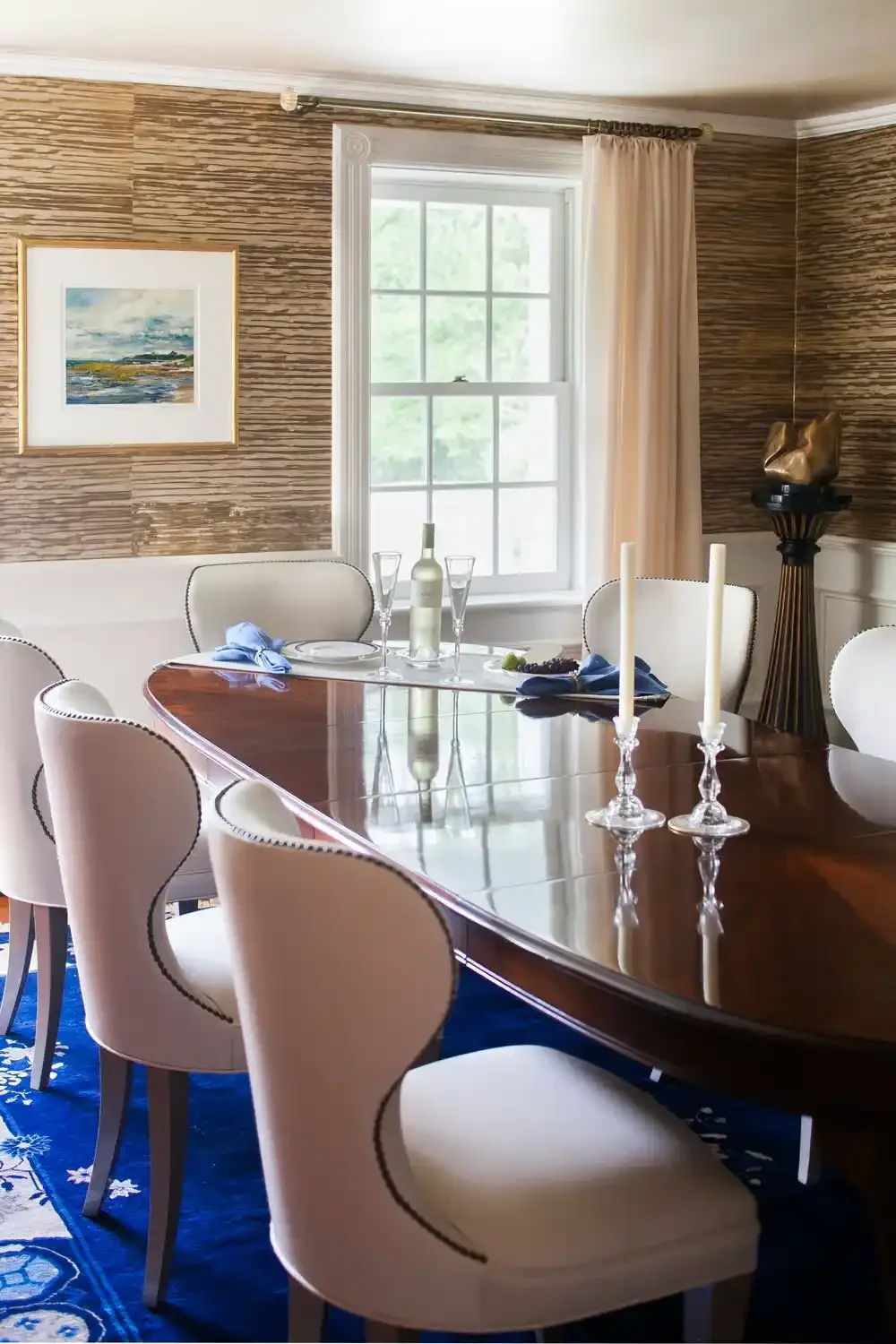 Dining room with a wooden table, white upholstered chairs, a window with cream curtains, and a table set with a bottle, glasses, and candles.