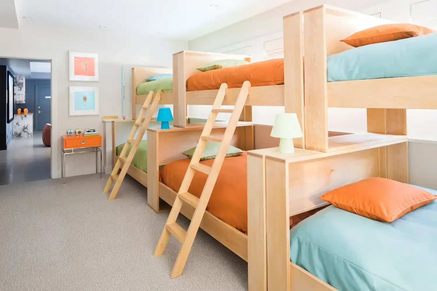 A room with four wooden bunk beds with colorful bedding and small ladders, bedside lamps, and artwork on the walls, with a doorway leading to another room.