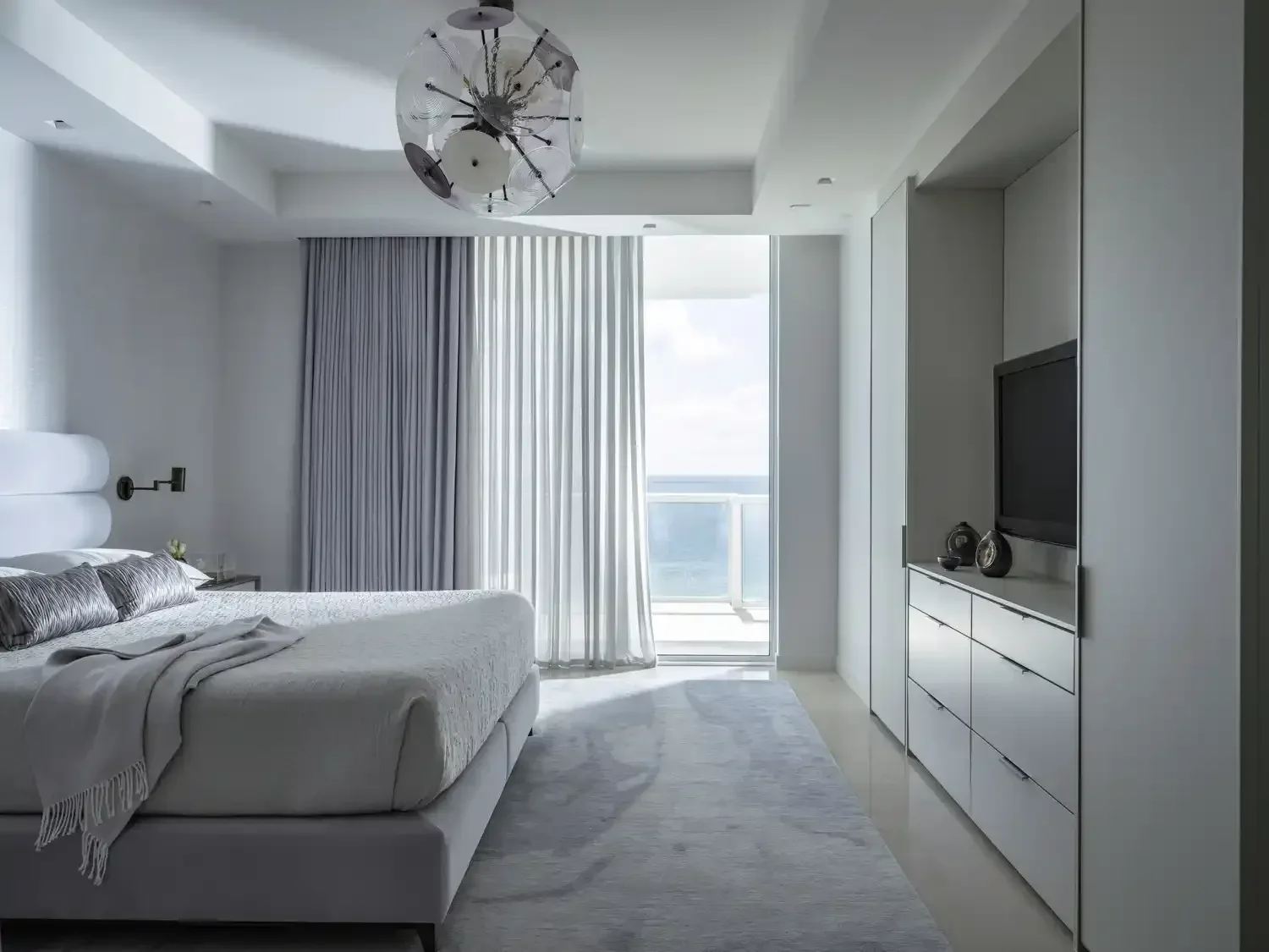 A modern bedroom with a large white bed, gray pillows and a blanket, a nightstand with a small plant, a wall-mounted black reading lamp, a flat-screen TV on a white dresser, light-colored curtains, a sliding glass door leading to a balcony, and a vie