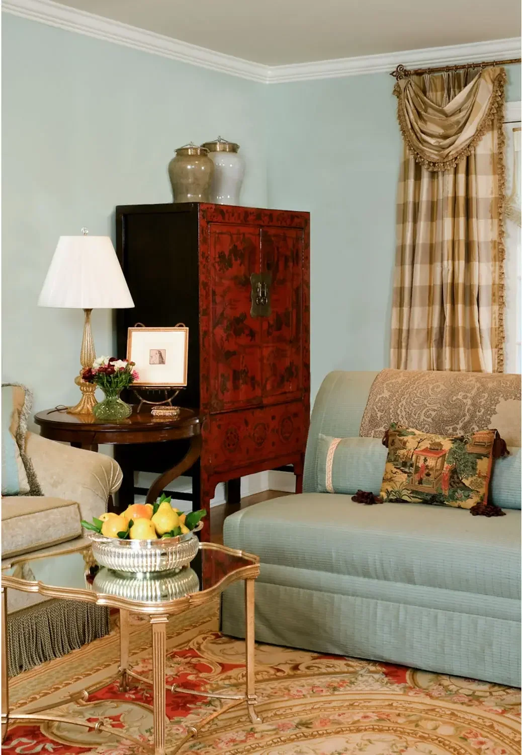 A cozy living room corner with light green walls, a patterned area rug, a blue sofa with a decorative pillow, a wooden side table with a lamp and flowers, a red decorative cabinet, and beige checkered curtains.