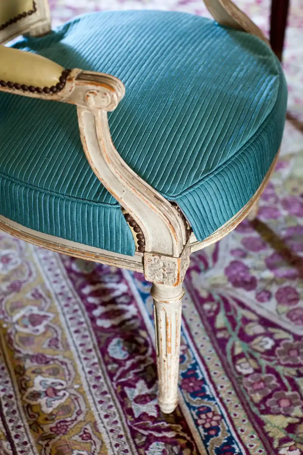 Close-up of a vintage chair with a distressed white wooden frame, teal velvet upholstery with vertical ribbing, on a patterned area rug.