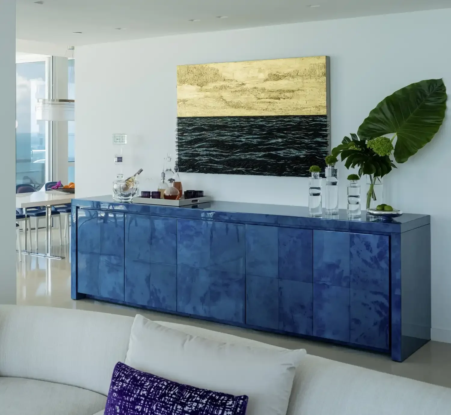 Modern interior with a blue marble sideboard, artwork with ocean and sky, large green plant, and decorative items.