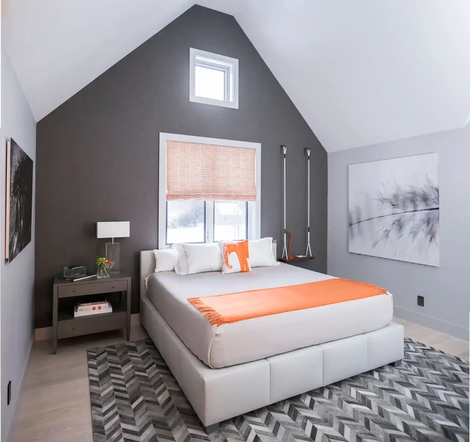 Modern bedroom with white bed, gray and black walls, orange accents, abstract wall art, window with orange and white shade, swinging chair, patterned area rug.