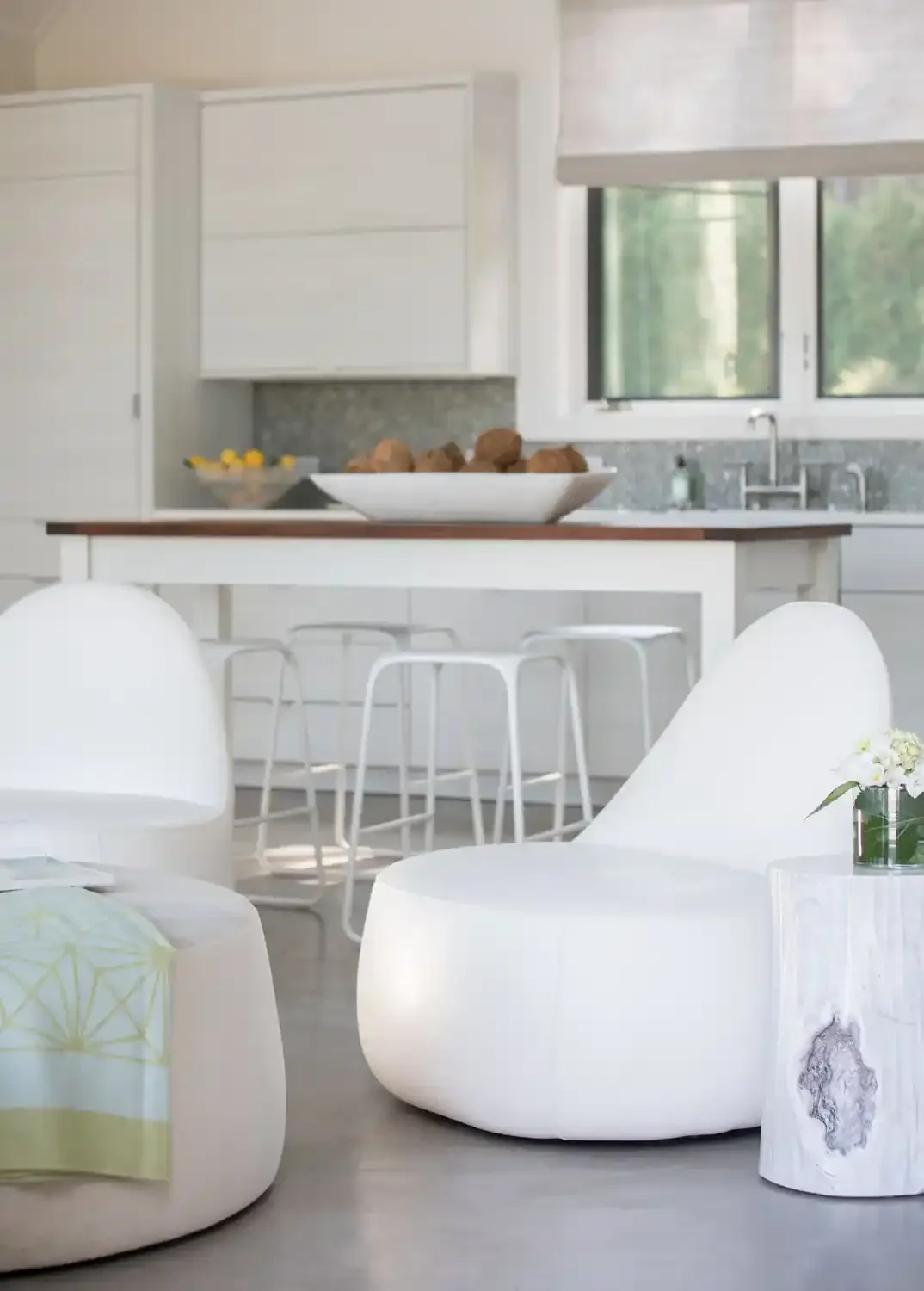 Modern kitchen with white cabinets, a large window, a countertop with a bowl of rocks, and a white dining area with stools.