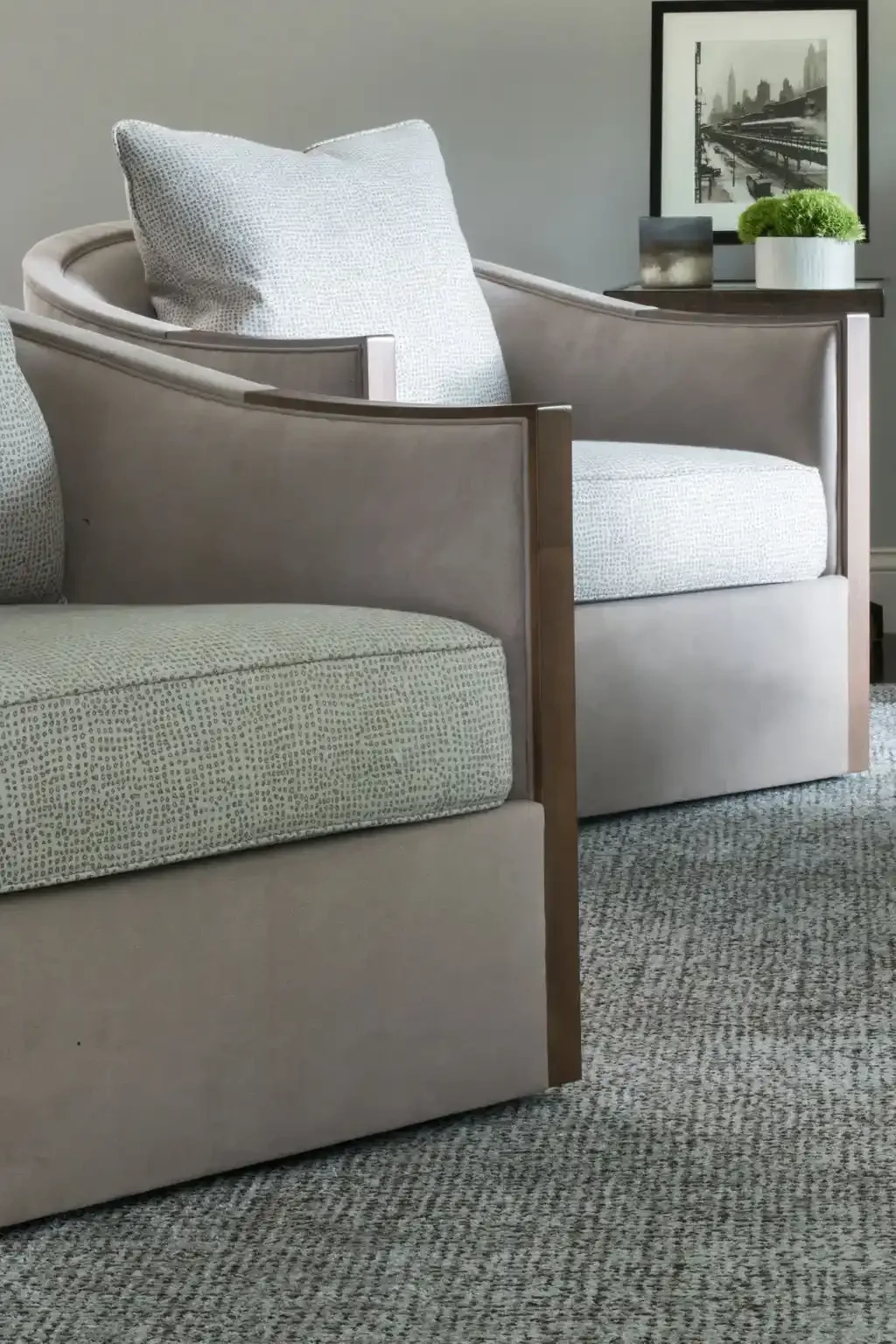 Close-up of a beige upholstered armchair with white textured cushions, in a living room with a gray wall, a wooden side table holding a framed black and white cityscape picture, a small metal object, and a white pot with green plants, with a patterne