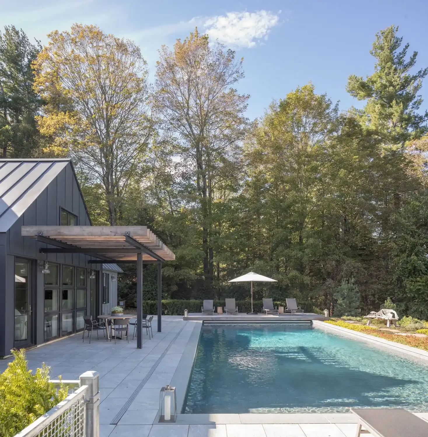 A modern house with large windows, a patio with a dining table and chairs, a swimming pool with lounge chairs and an umbrella, and lush trees in the background on a sunny day.