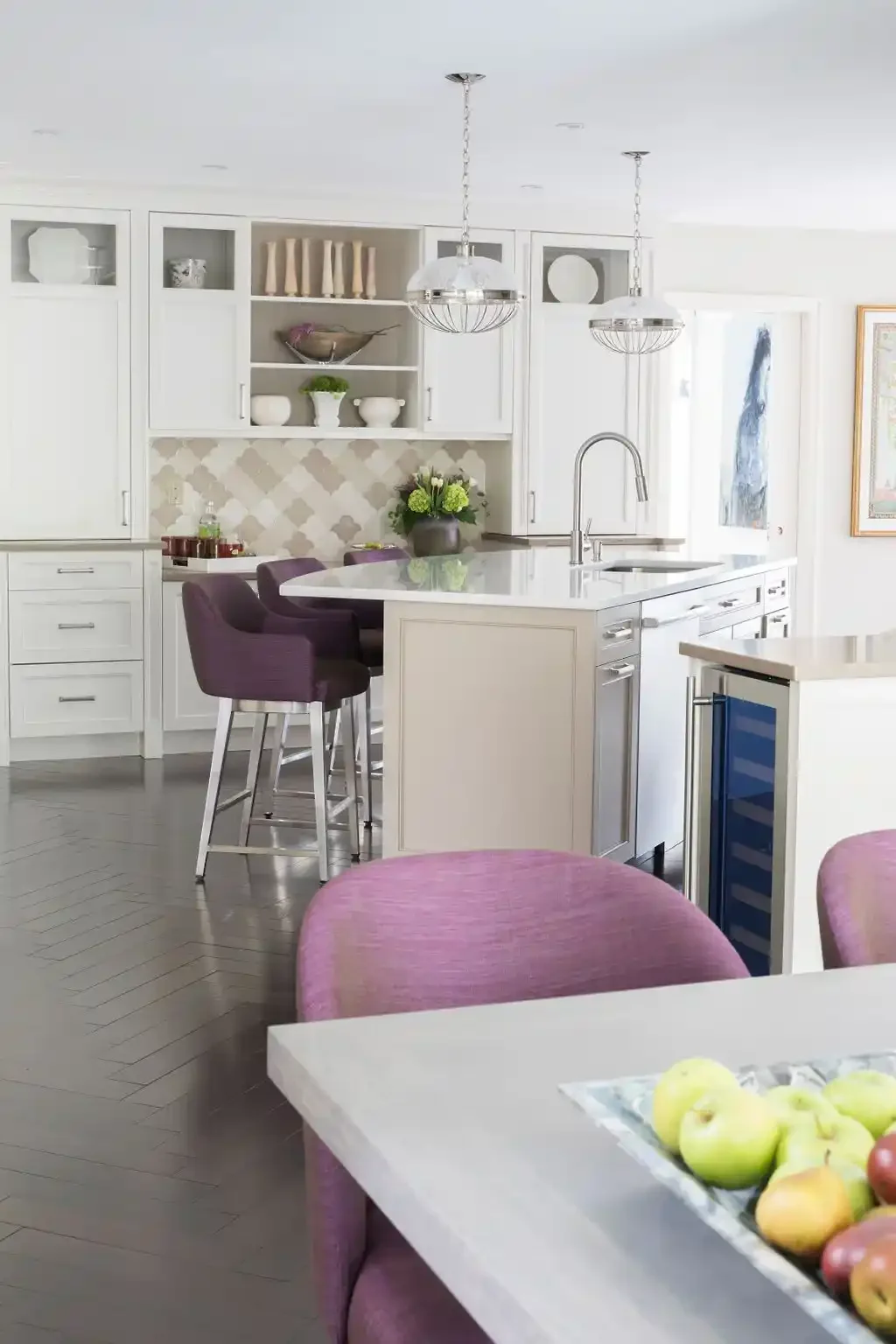 Modern white kitchen with purple chairs around a marble island, open shelving with decorative dishes, a nearby dining table with apples, and artwork on the walls.