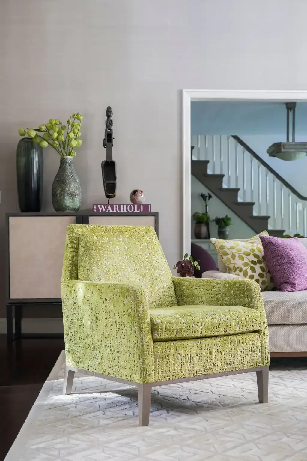 Bright green mid-century armchair in a modern living room with decorative pillows and a side table with vases and books, staircase visible through doorway.