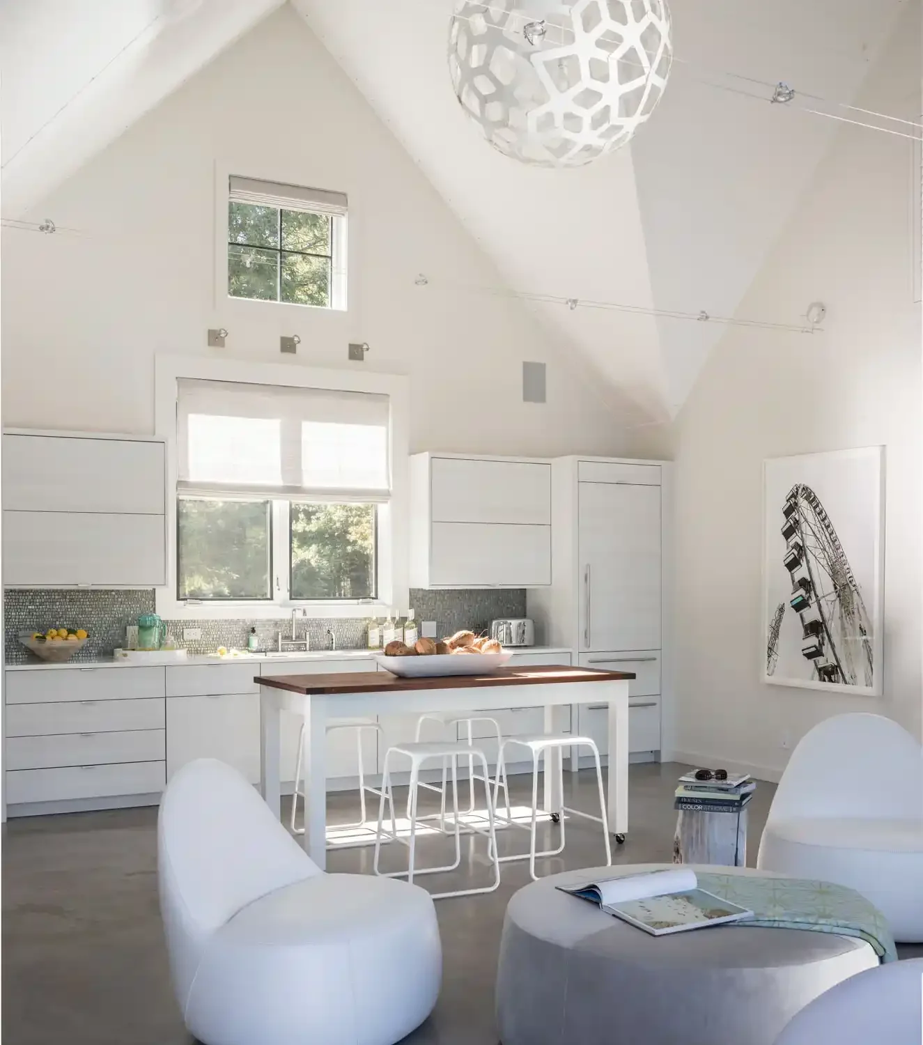 Bright, minimalist kitchen with white cabinets, a wooden kitchen island, and large windows. Modern decor includes a framed Ferris wheel artwork and a geometric hanging light fixture.