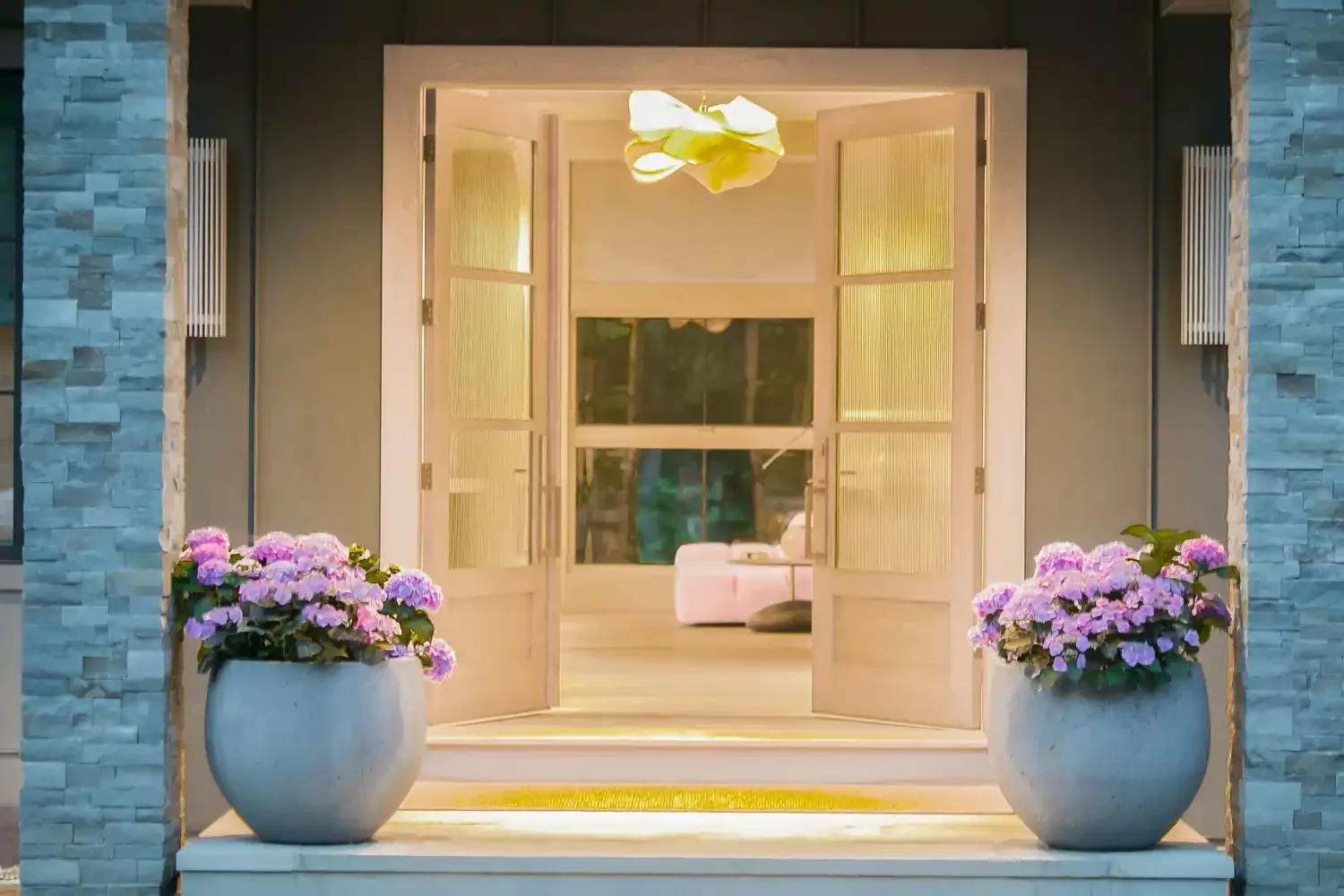 Open front door of a house with two large flower pots with pink and purple flowers on each side of the entrance, revealing a cozy indoor space with a pink sofa inside.
