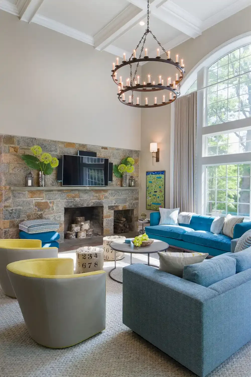 Living room with a stone fireplace, a large window, a chandelier hangings from a white ceiling, a flat screen TV above the fireplace, and various colorful sofas and chairs.