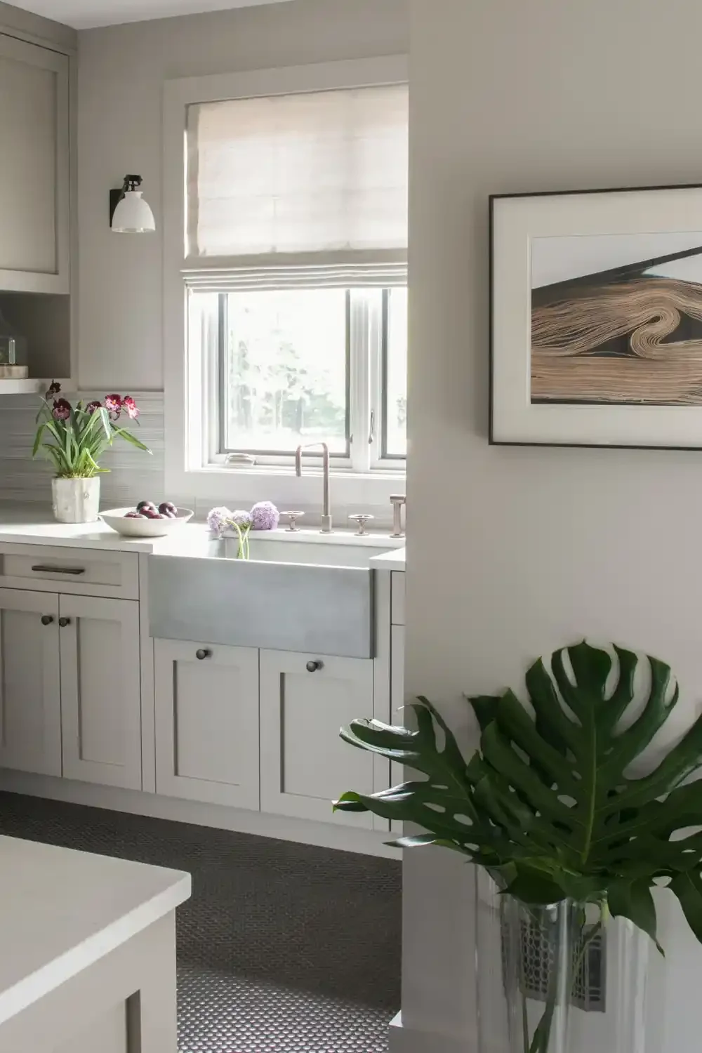 Bright kitchen with white cabinets, a farmhouse sink, and a window above it. There is a potted plant with pink flowers on the counter and a framed abstract art on the wall. A large green leaf plant is in the foreground.