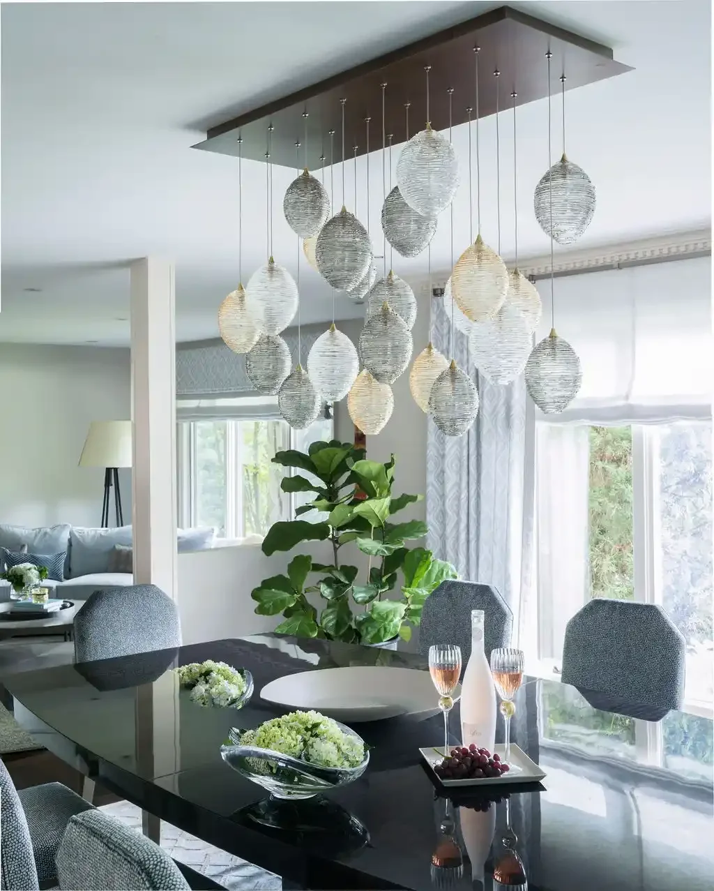 Modern dining room with a black table, gray chairs, a large green plant, and a hanging light fixture with multiple white and gray oval-shaped lights.