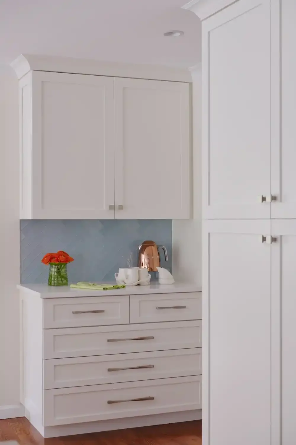 A white kitchen cabinet with drawers and upper cabinets, a small countertop with a copper kettle, white cups, a small white appliance, a vase with orange flowers, and a lime green cloth.