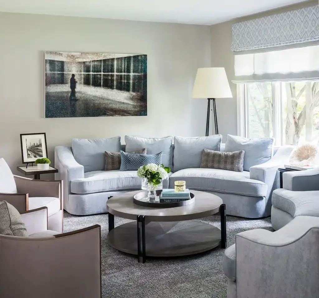 Bright living room with a large gray sectional sofa, patterned throw pillows, a round coffee table with books and a flower arrangement, a floor lamp, and window with curtains allowing sunlight in.