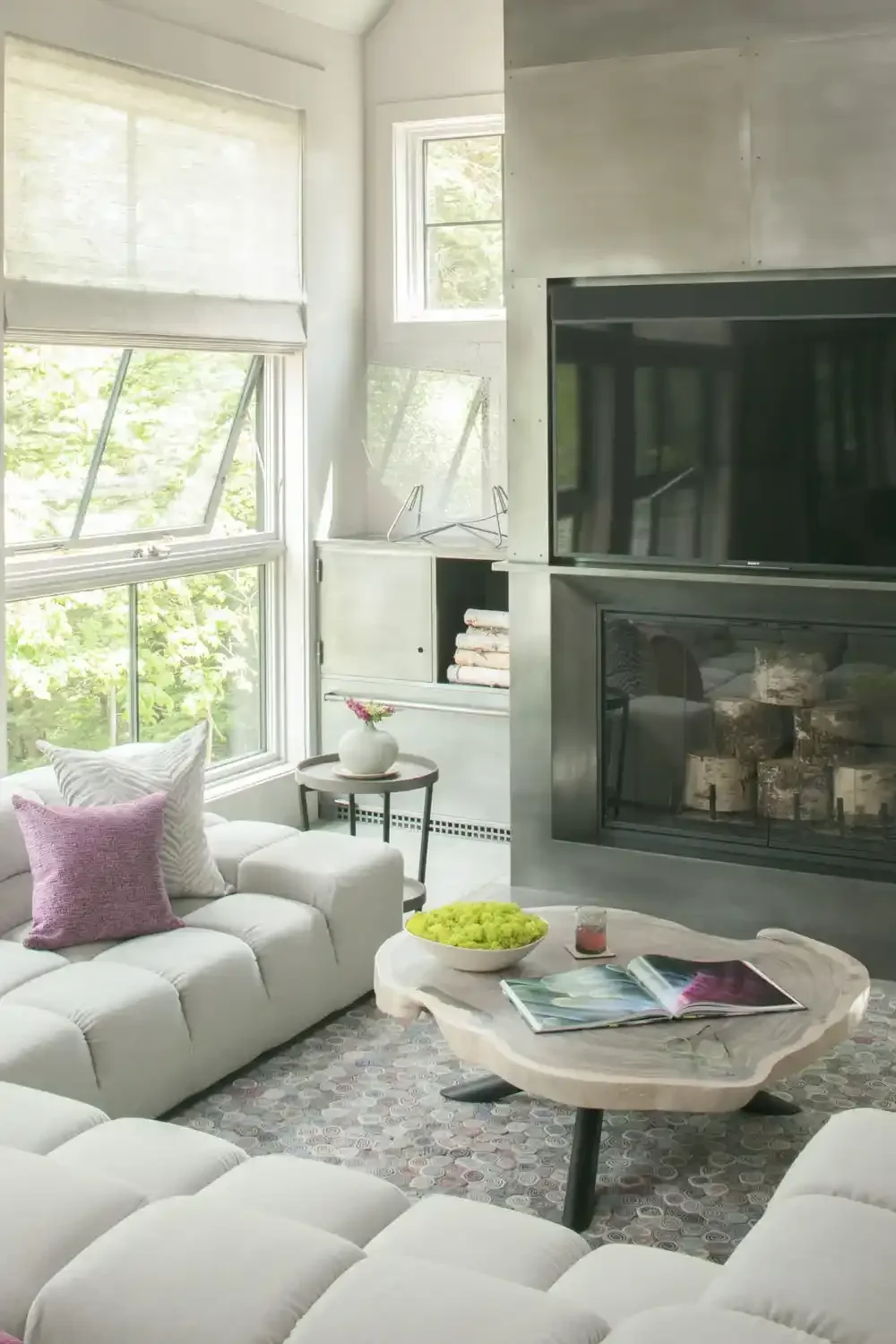 Bright living room with large windows, white sofa with pink and striped pillows, a round coffee table with magazine and a bowl, a small side table with a flower vase, a fireplace, and a wall-mounted TV.