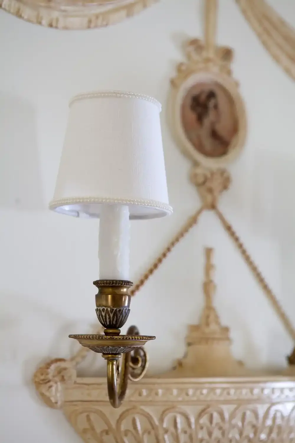 Close-up of a vintage wall sconce with a white fabric lampshade, mounted on an ornate cream-colored wall with decorative moldings and a framed portrait in the background.