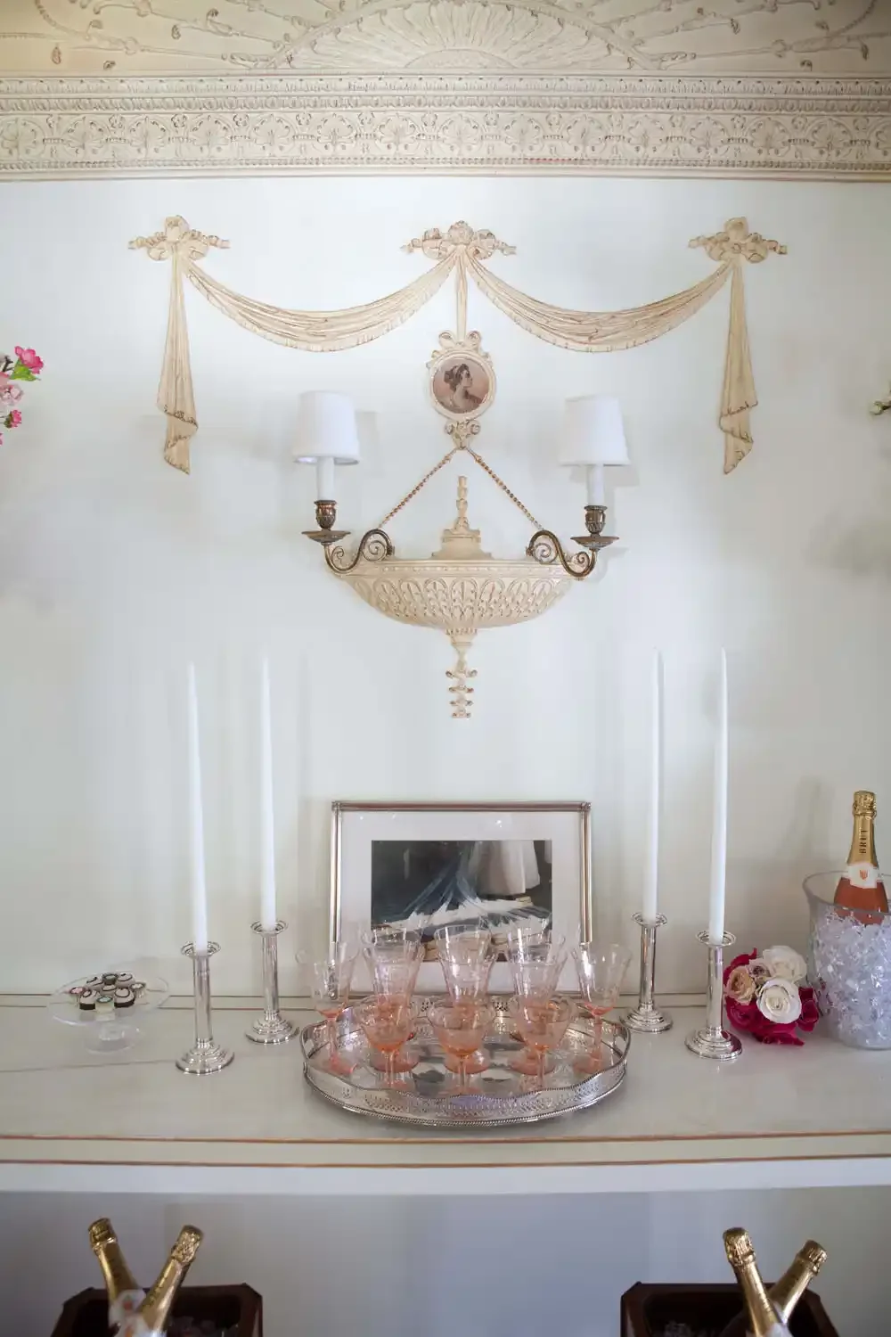 Vintage ornate wall sconce with two white candles, a tray of pink glasses, flowers, and a bottle of champagne on a white surface, with decorative molding and draped fabric on the wall behind.