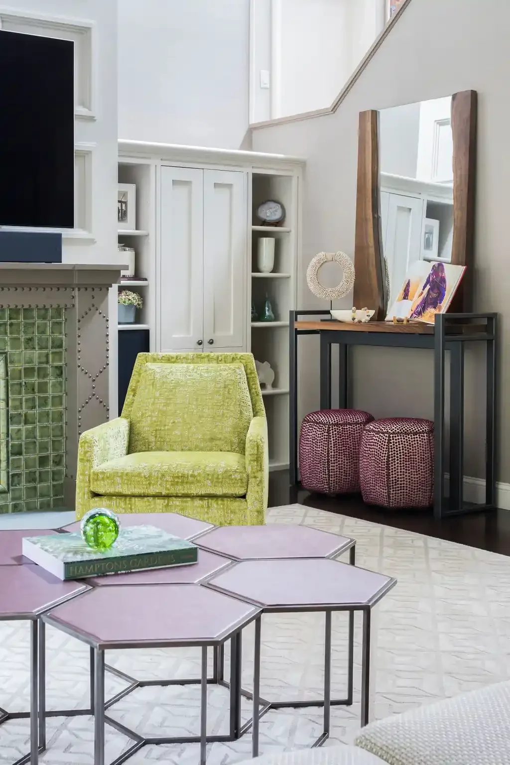 Living room with green armchair, hexagonal pink tables, white rug, decorative shelves, mirror, and artwork.