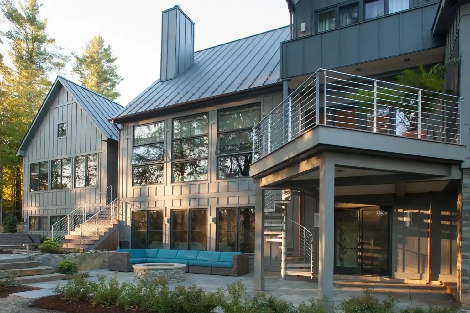 Modern multi-story house with large windows, metal roof, and outdoor balcony. There's a garden with a curved outdoor seating area and fire pit.