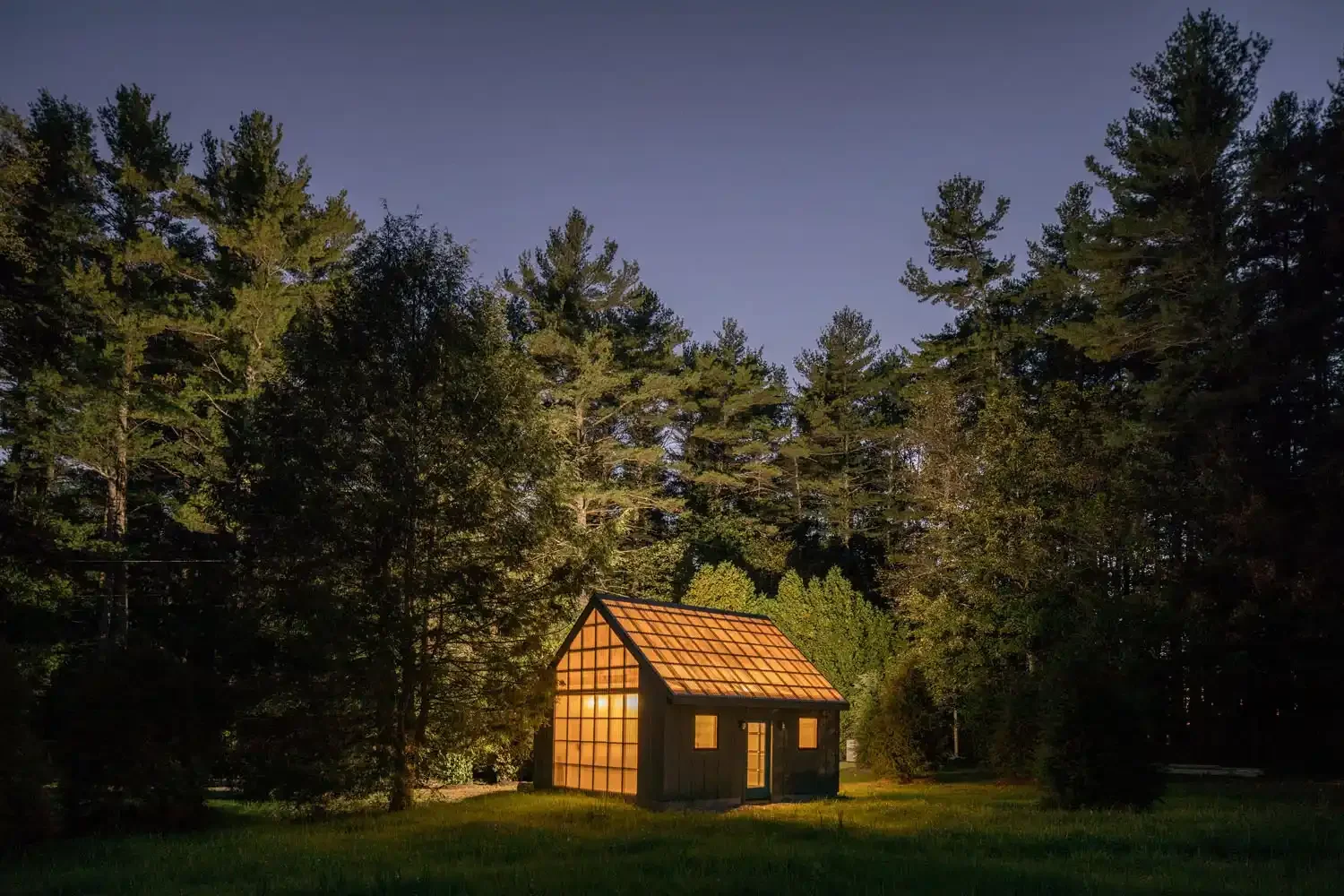 A small wooden house with large windows glowing warmly, situated on green grass, surrounded by tall pine trees under a clear evening sky.