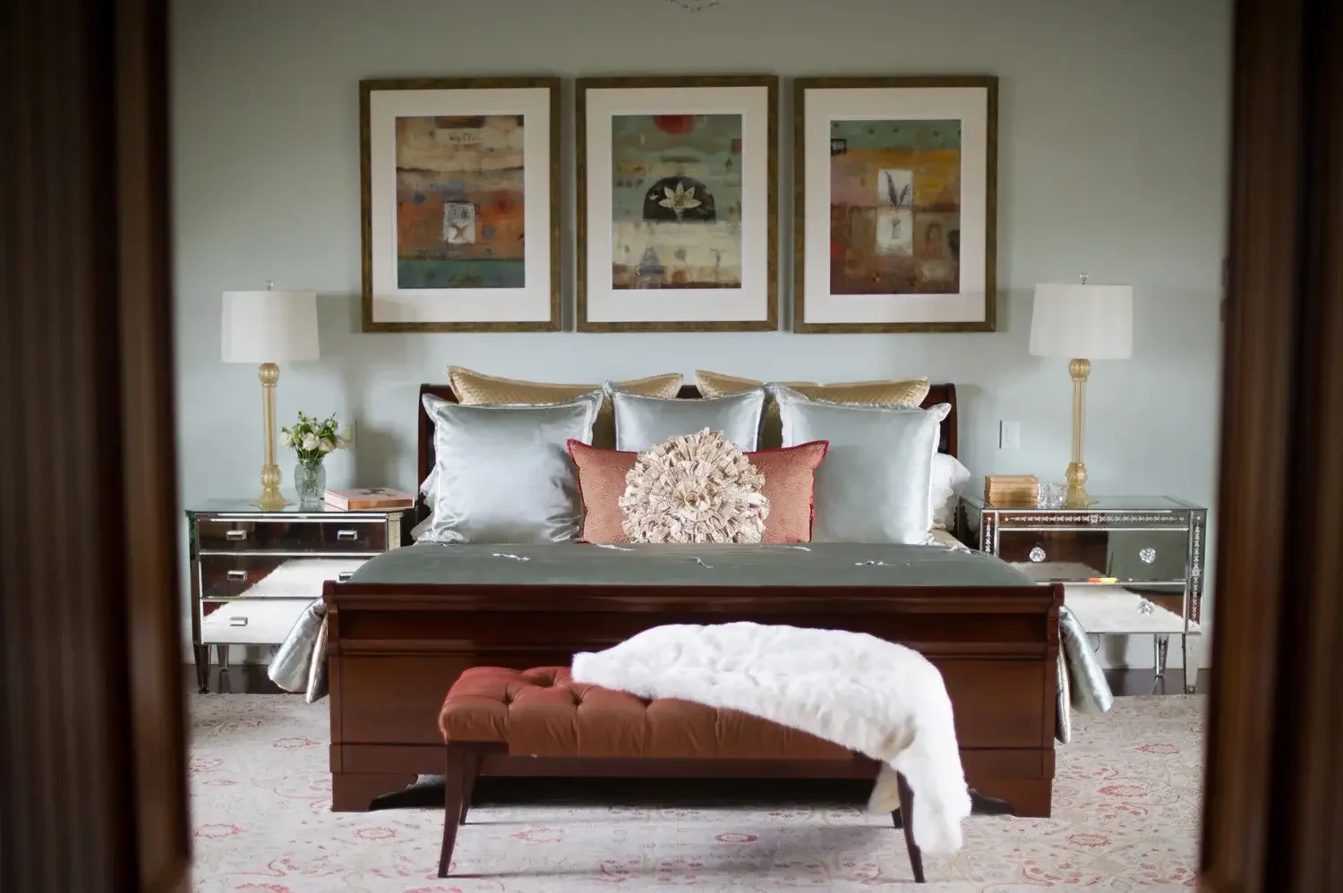 Bedroom with a wooden bed, decorative pillows, a bench with a white throw, two mirrored nightstands with lamps, and framed artwork above the bed.