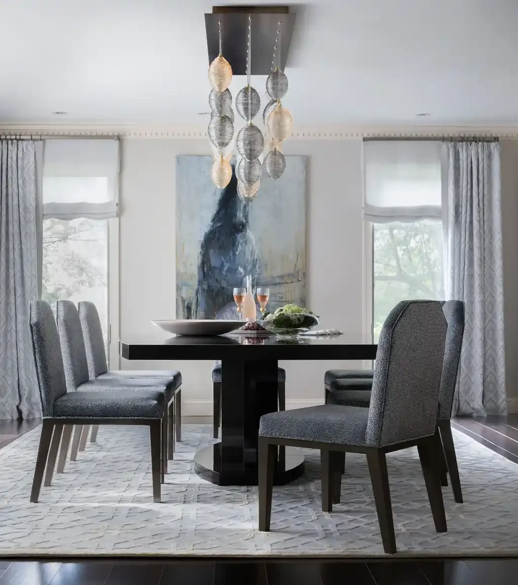 Modern dining room with six gray upholstered chairs around a black round table, which has a large white bowl, two glasses of pink drinks, and a green plant on it. There are two large windows with white curtains, a gray abstract painting on the wall, 