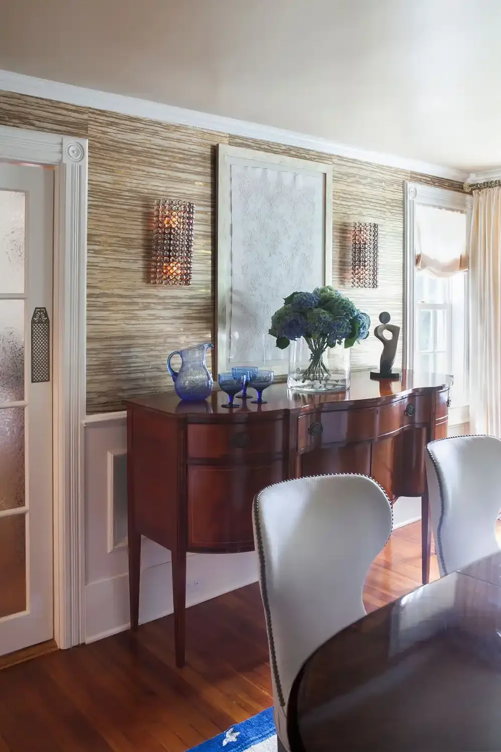 A dining room with a wooden sideboard decorated with blue glassware, a glass vase of blue and green hydrangeas, and a modern sculpture. The wall behind features textured wallpaper with two decorative light fixtures. There are white upholstered chairs