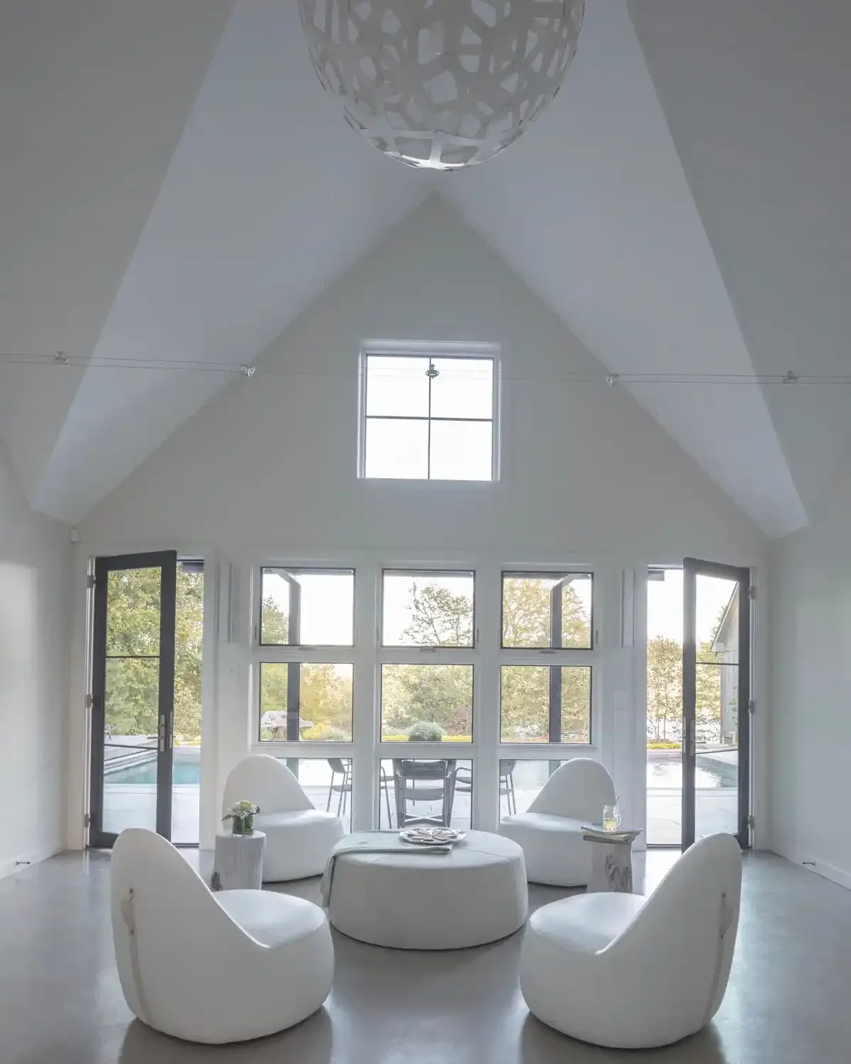 Bright, modern living room with large windows and white furniture, including a circular coffee table and four armchairs, in a space with a high, vaulted ceiling and a geometric pendant light.
