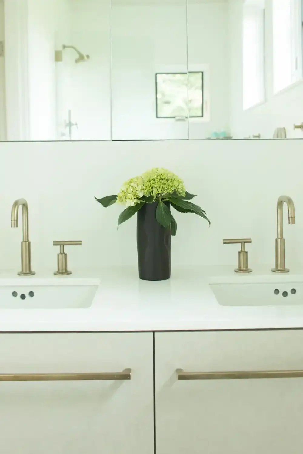 A double sink bathroom vanity with gold faucets, a black vase with green and white hydrangea flowers, white cabinets, and a mirror with a reflection of a window and shower.
