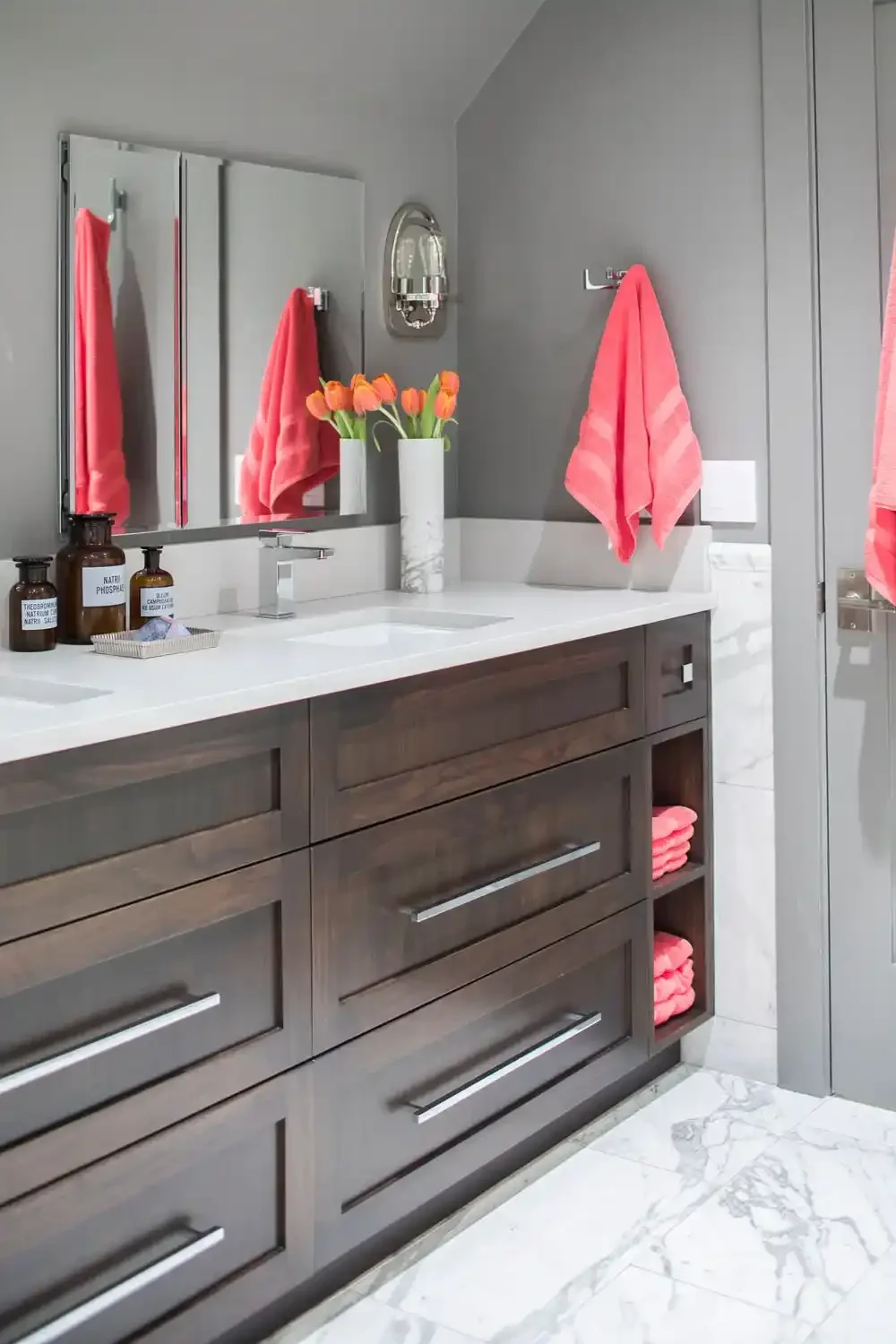 Modern bathroom with dark wooden vanity, white marble countertop, pink towels, and decorative orange tulips in a white vase.