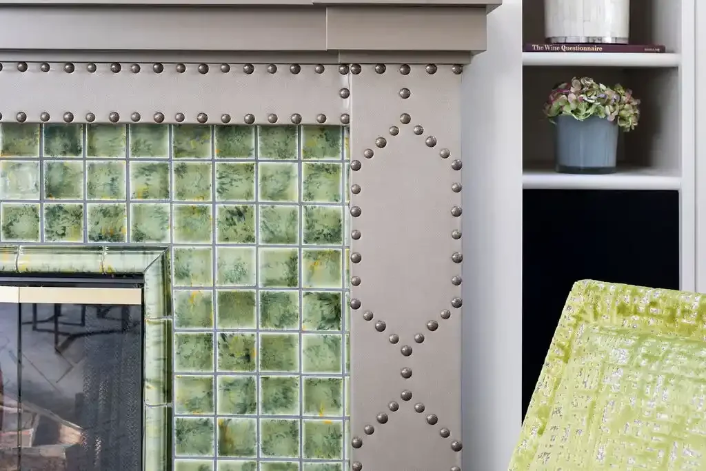Close-up of a fireplace with a border of silver decorative studs, surrounded by green tile and a white mantel, with a shelf holding a potted plant and books in the background.