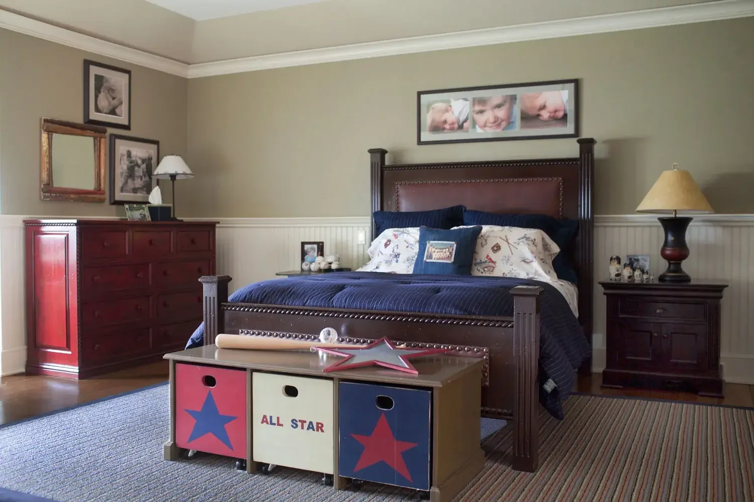 A bedroom with a large wooden bed, navy blue bedding, and decorative pillows. There are wooden nightstands with lamps on each side of the bed. A colorful storage cube with star designs and the words 'ALL STAR' is at the foot of the bed. Framed family