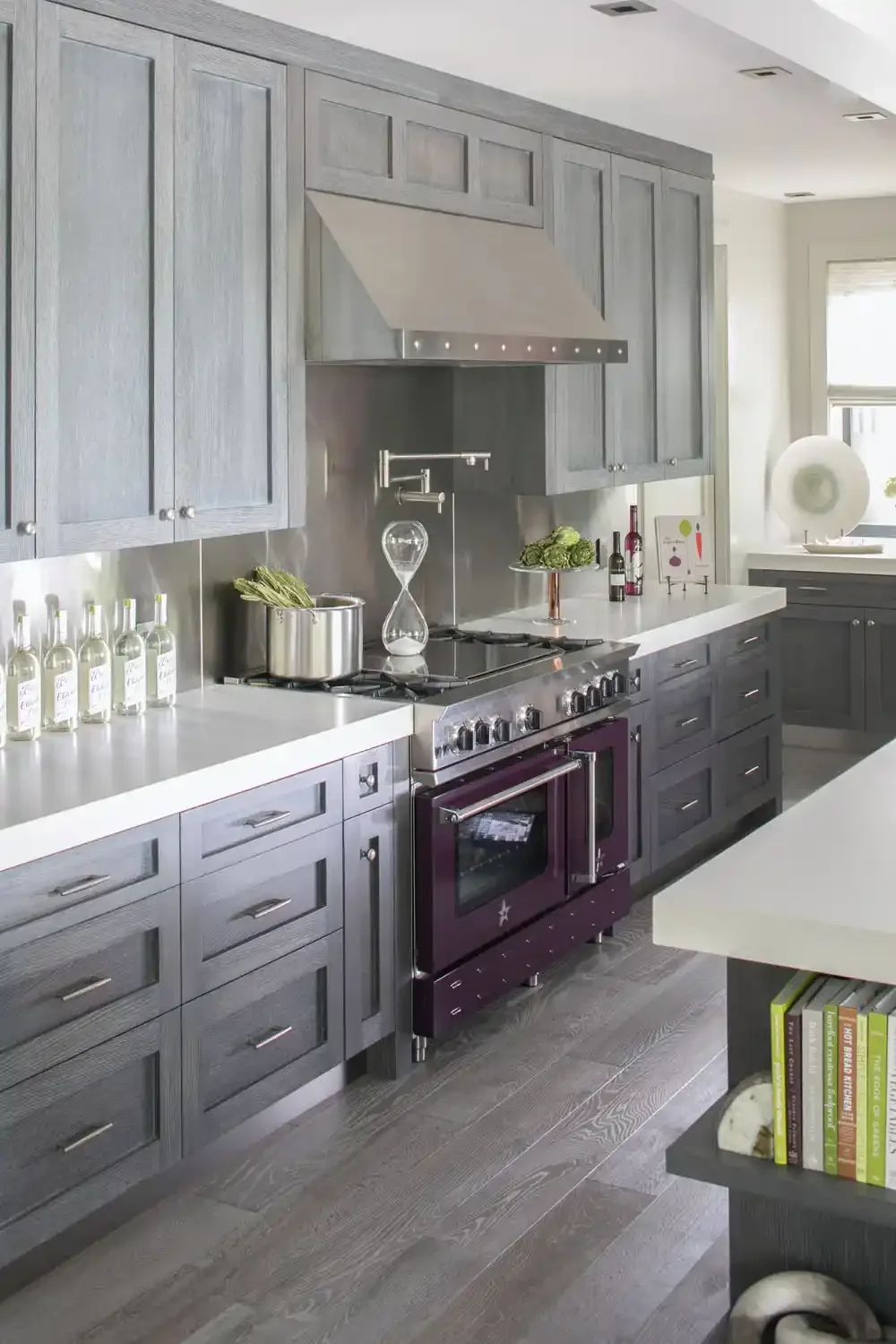 Modern kitchen with gray cabinets, white countertops, and stainless steel appliances, including a purple oven. Kitchen decor includes a glass hourglass, bottles of olive oil, a potted herb, and a small framed artwork. There is a window with a roll-up