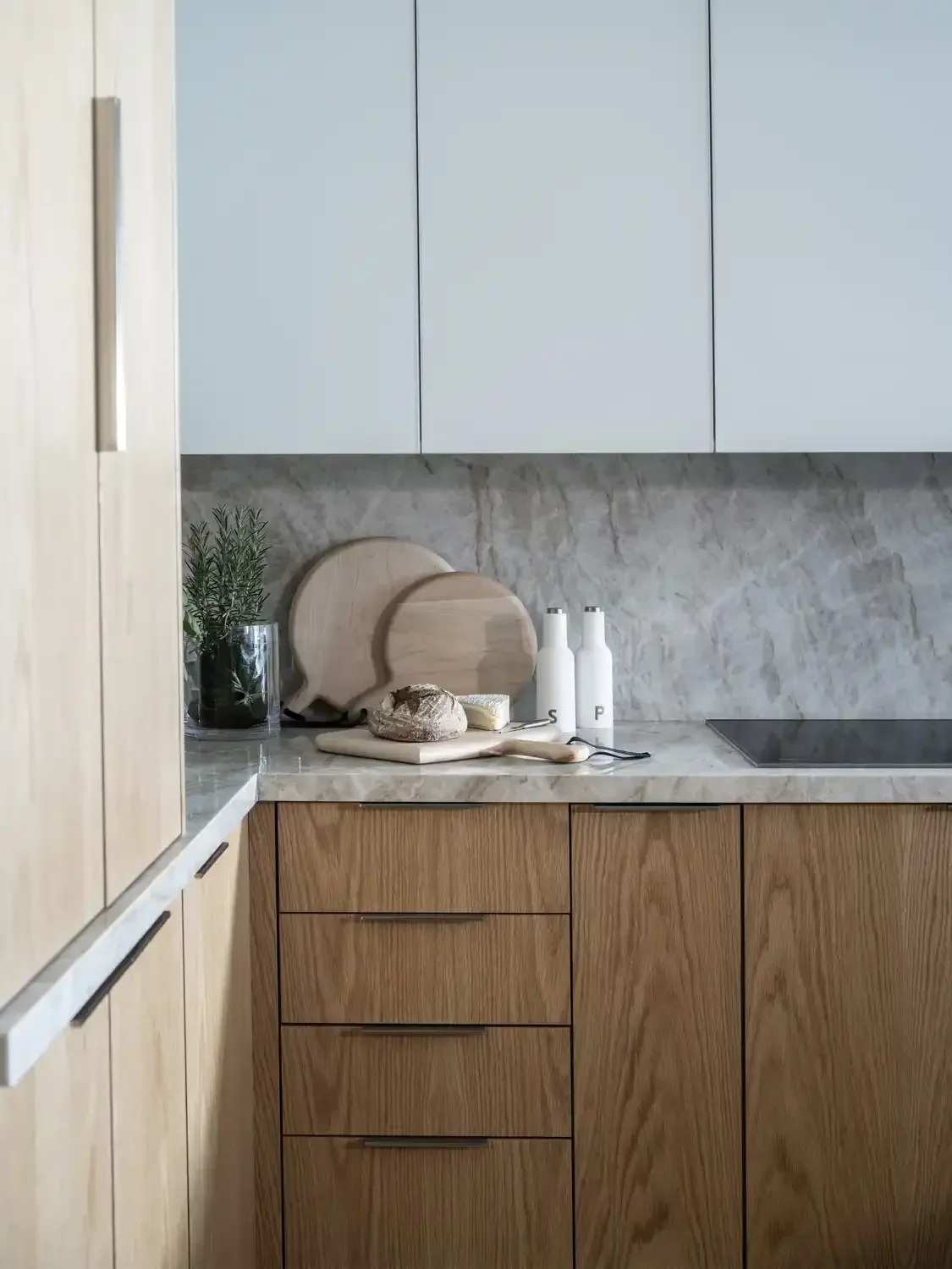 Modern kitchen with light wood lower cabinets, abstract light blue upper cabinets, marble backsplash and countertop, and various kitchen items including two wooden cutting boards, a potted herb, and white bottles.