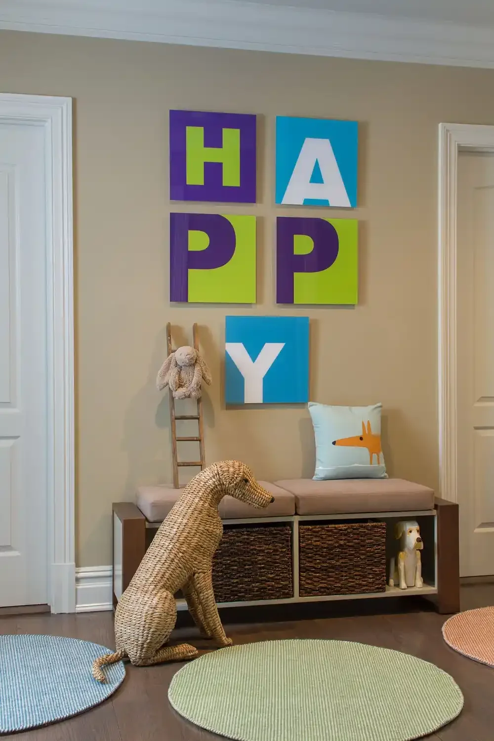 Decorative wall art spelling out 'HAPPY' with colorful blocks in a child's playroom. A woven dog sculpture is seated on the floor, with a lounge chair featuring a pillow with a fox illustration. A small stuffed animal hangs on a ladder leaning agains
