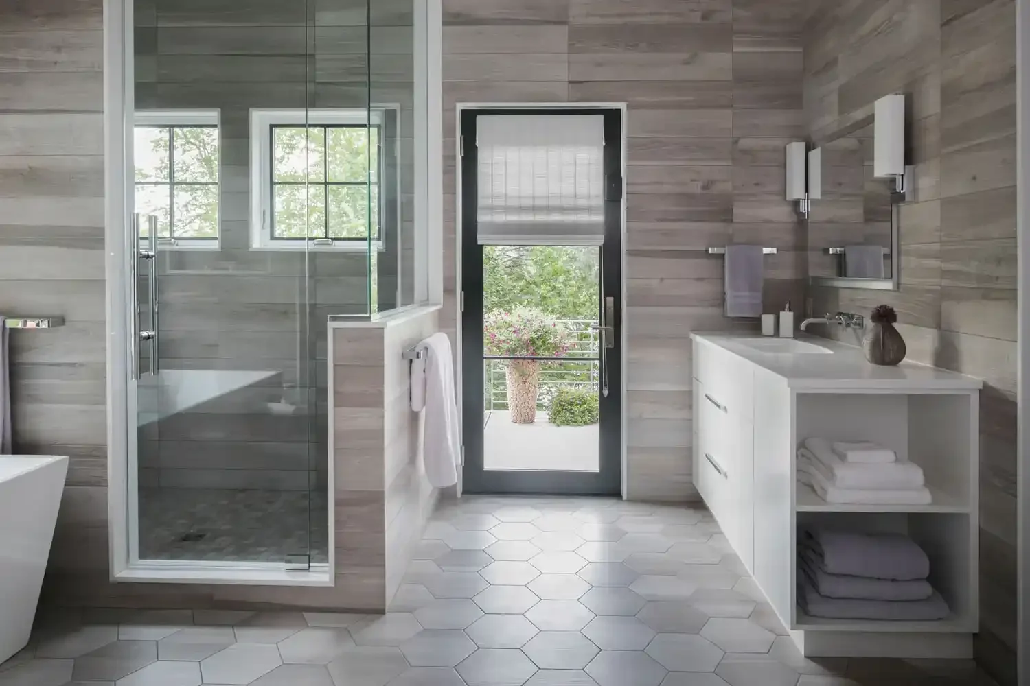 Modern bathroom with gray wood-paneled walls, white hexagonal floor tiles, a glass-enclosed shower, a white vanity with towels, and a door leading to a balcony with greenery outside.
