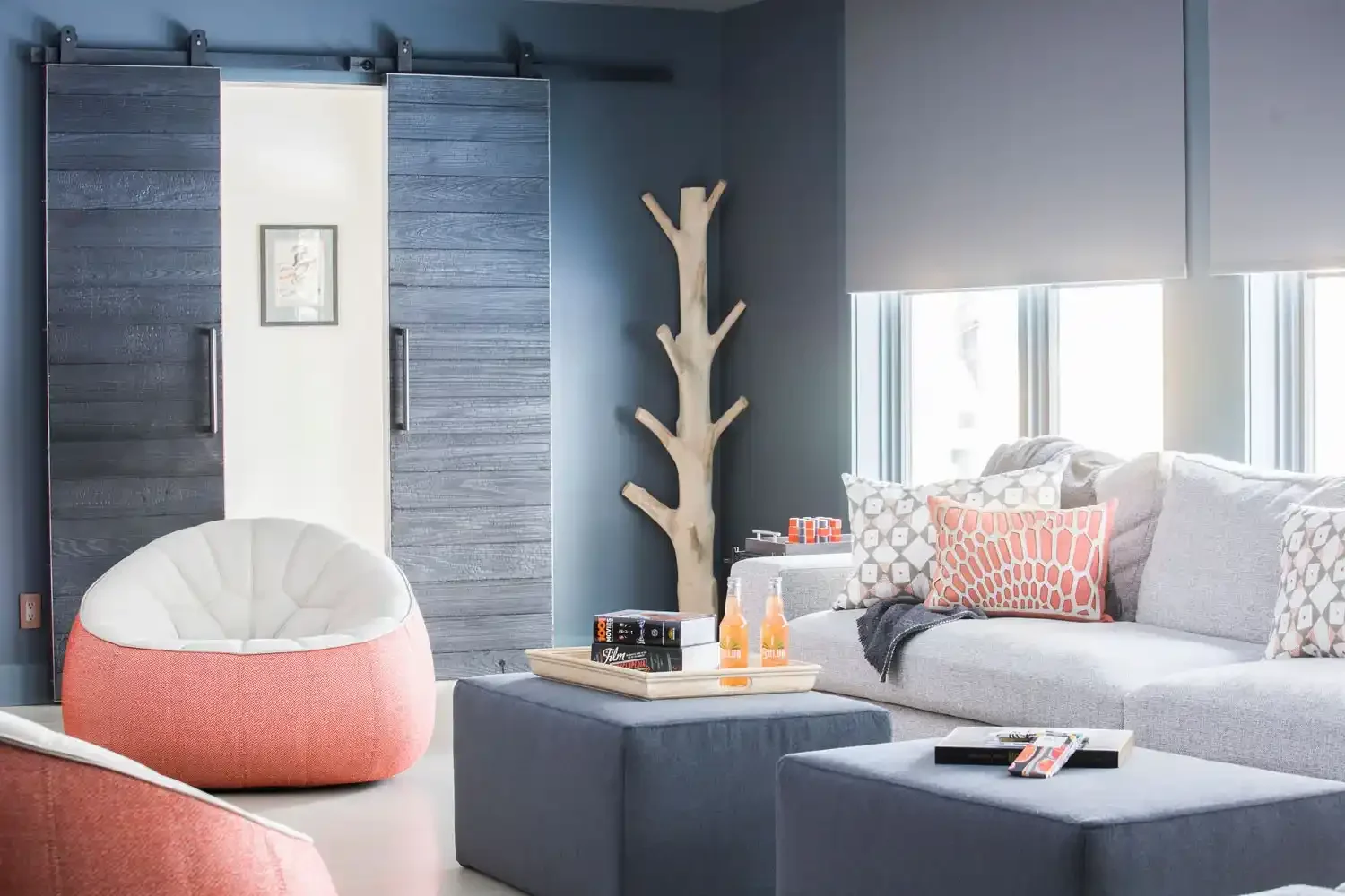 Living room with a large gray sectional sofa, various patterned pillows, a cozy round chair with pink and white fabric, a blue ottoman with books and drinks, a tall wooden branch-shaped coat rack, and windows with blinds.