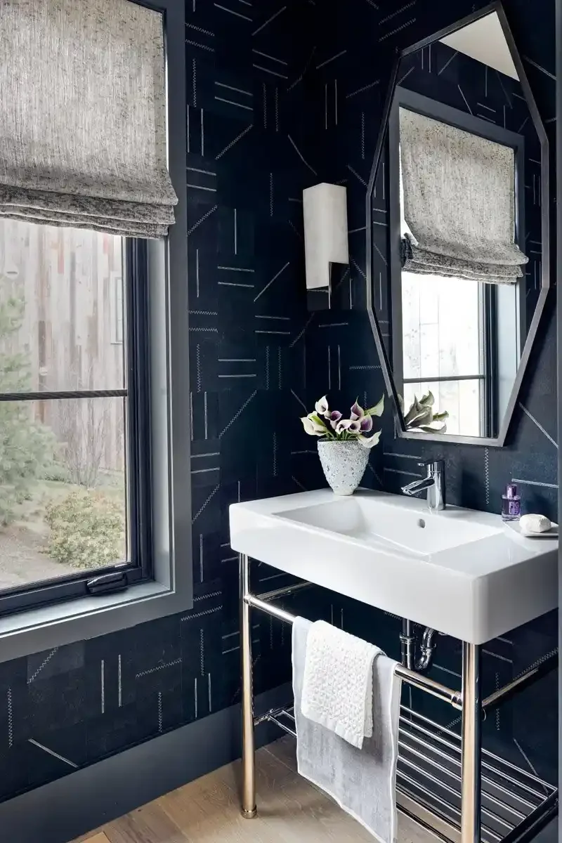Modern bathroom with a rectangular white sink, a large angular mirror, a window with a gray Roman shade, a small white wall-mounted light fixture, a towel hanging on a metal towel rack, and a vase with flowers on the sink. The walls are dark with a g