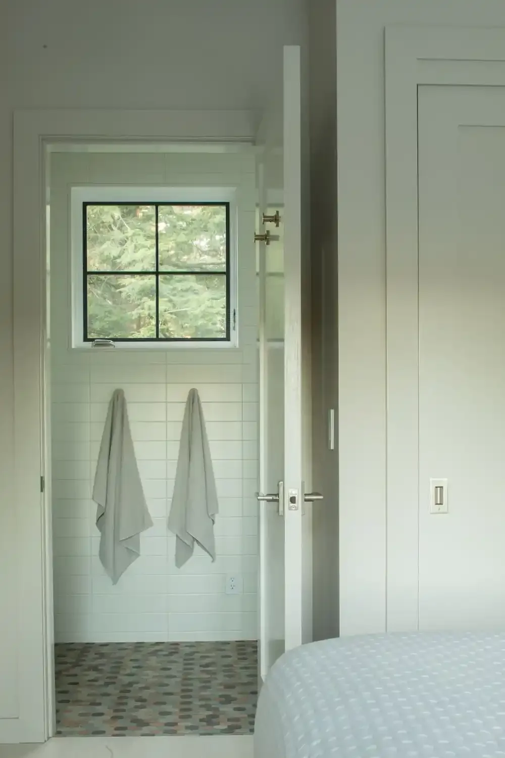 A doorway leading to a small outdoor space with two white towels hanging on hooks, a window showing trees outside, and part of a bed in the foreground.